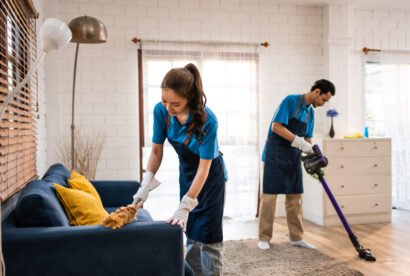 Asian young man and woman cleaning service worker work in living room. Attractive housekeeper cleaner team wear apron and cleaning messy dirty floor for housekeeping housework and chores in house.
